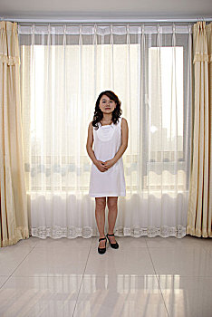 一名年轻女性穿着白色的连衣裙站在挂着白色窗帘的窗户前