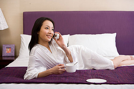 一个女人趴在床上边打电话边喝咖啡