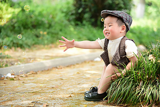 阳光下林荫小路上穿着短袖西服坐在草坪上伸手抓泡泡的小男孩