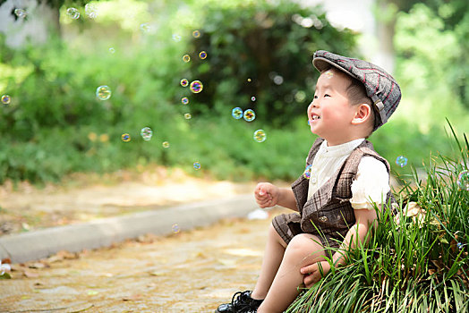 阳光下林荫小路上穿着短袖西服坐在草坪上看着泡泡开心的小男孩