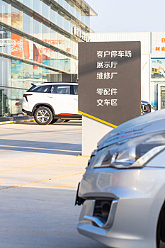 一家汽车4s店外摆放着的新车旁边立着一个指示牌,上面写着停车场,交车区等信息