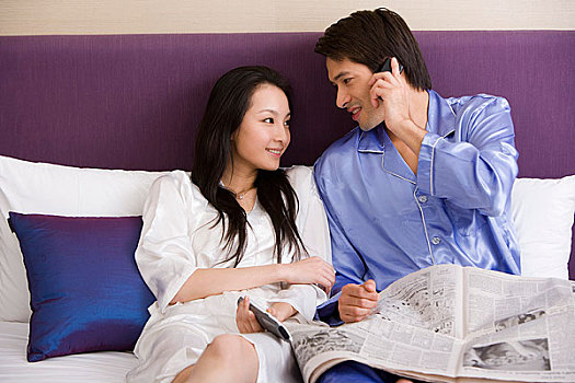 一对年轻的夫妻坐在床上,男的看报接电话,女人看电视