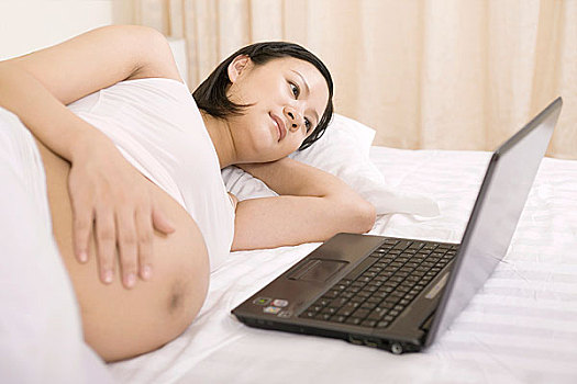 对着笔记本电脑侧卧在床上的孕妇