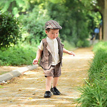 阳光下林荫小路上穿着短袖西服手拿树枝玩耍的小男孩