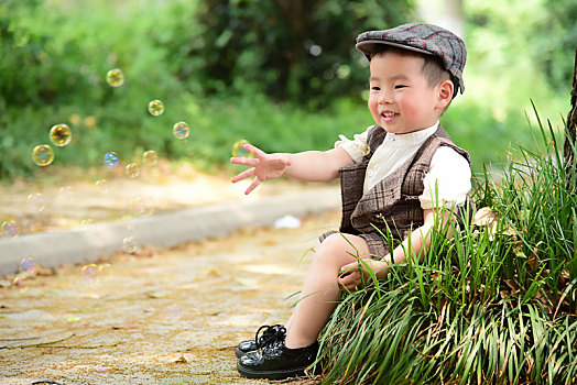阳光下林荫小路上穿着短袖西服坐在草坪上伸手抓泡泡的小男孩