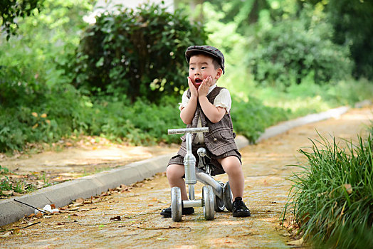 阳光下林荫小路上穿着短袖西服坐在滑滑车上双手托下巴小男孩