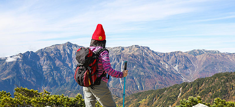一个手拿登山杖站在山顶看着远方风景的女性徒步旅行者背影