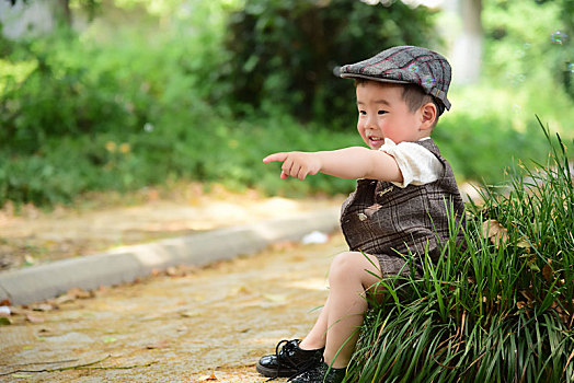阳光下林荫小路上穿着短袖西服坐在草坪上伸手指着前方的小男孩