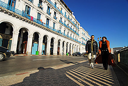 algeria,alger,couple,walking,on,sidewalk,while,car,moving,street,by,residential,structure