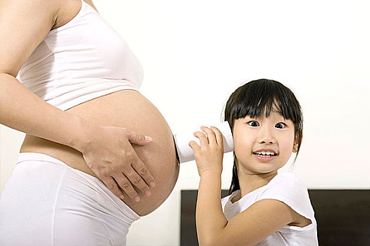 坐在床上用纸筒听怀孕的妈妈腹部的小女孩