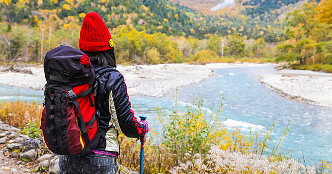 一个手拿登山杖站在秋天山谷河边的女性徒步旅行者背影