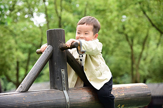 林荫下坐在树杆翘翘板上玩耍开心的小男孩
