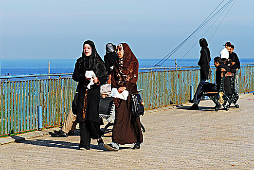 algeria,alger,side,view,of,girls,wearing,burka,walking,by,railing,while,couples,sitting,in,background,sea