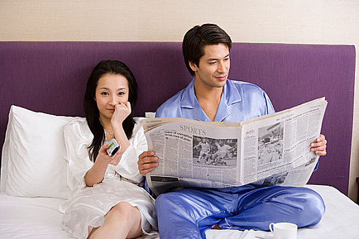 一对年轻的夫妻坐在床上看报纸和电视