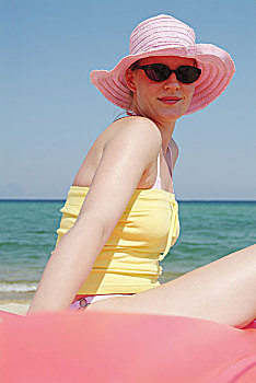 女人,年轻,太阳镜,帽子,气垫,海滩,坐,侧面,人,20-30岁,头饰,草帽,太阳帽,粉色,上面,黄色,晴朗,看镜头,户外,夏天,复原,休闲,放松,轻松,度假