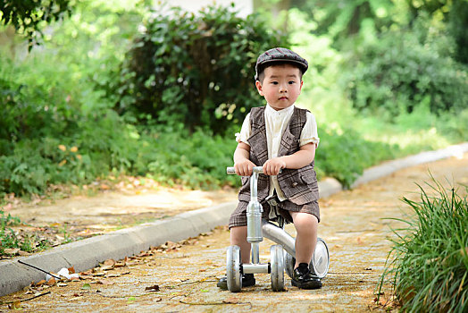 阳光下林荫小路上穿着短袖西服坐在滑滑车上开心的小男孩