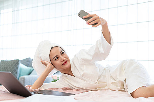 年轻女子穿着浴袍在床上使用手机自拍