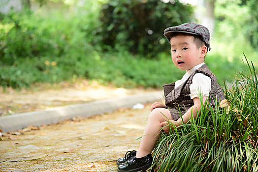 阳光下林荫小路上穿着短袖西服坐在草坪上玩泡泡的小男孩