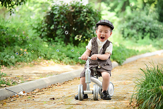 阳光下林荫小路上穿着短袖西服坐在滑滑车上闭眼大笑的小男孩