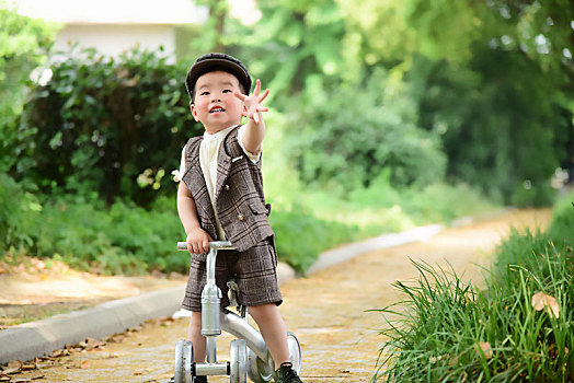 阳光下林荫小路上穿着短袖西服扶着滑滑车伸手抓泡泡的小男孩