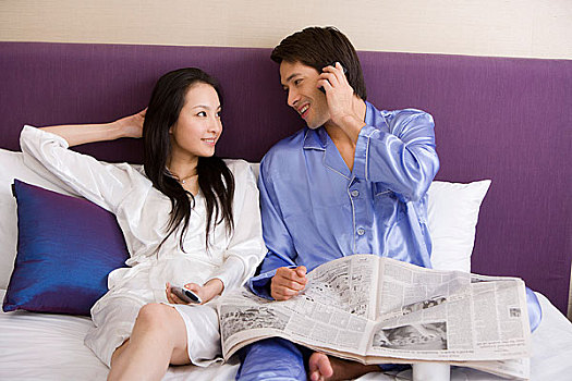 一对年轻的夫妻坐在床上,男的看报接电话,女人看电视