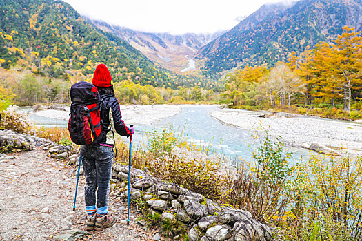一个手拿登山杖站在秋天山谷河边的女性徒步旅行者背影