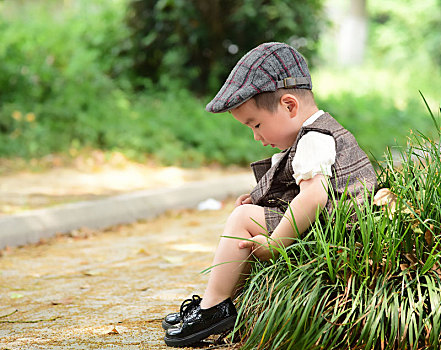 阳光下林荫小路上穿着短袖西服坐在草坪上低头挠腿的小男孩
