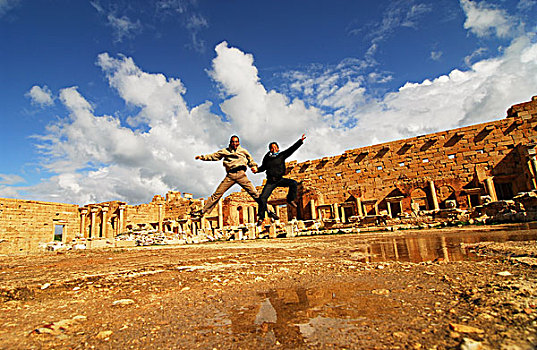 libya,leptis,magna,couple,jumping,in,the,air,with,ruins,background,unesco,world,heritage,site