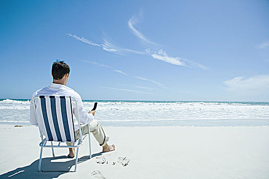 男人,坐,折叠椅,海滩,拿着,手机,后视图
