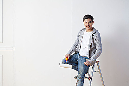 坐在梯子上准备粉刷墙壁的青年男子