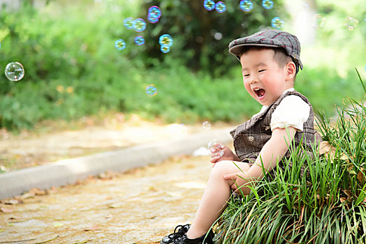阳光下林荫小路上穿着短袖西服坐在草坪上玩泡泡的小男孩