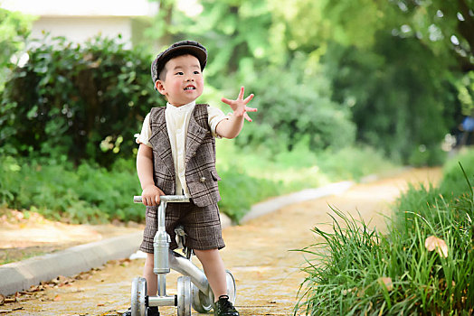 阳光下林荫小路上穿着短袖西服扶着滑滑车伸手抓泡泡的小男孩