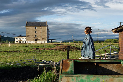 草原上一个穿牛仔的女孩站在生锈的铁箱旁回望远方的房子